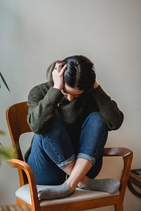 Woman sitting on a sofa with head in hands, showing symptoms of cervicogenic headache linked to neck dysfunction and poor posture.
